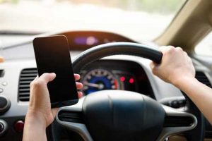 vehicle interior shows a driver compartment and driver's arms. One hand on the steering wheel and one hand holding a cell phone.