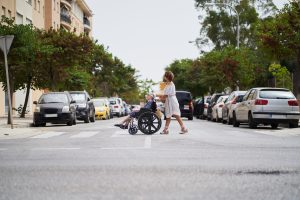 Two Pedestrians crossing an intersection at a crosswalk. One of the Pedestrians is seated in a wheelchair. 