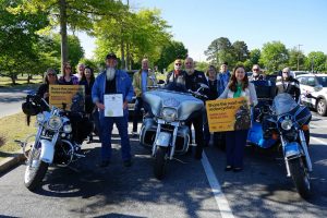 A group of motorcycle riders, safety advocates, and transportation officials stand in a parking lot between three large touring motorcycles during a motorcycle safety awareness event. Two participants hold signs reading “Share the road with motorcyclists – Bikers Have Families Too,” while another holds an official proclamation. Trees and parked vehicles are visible in the background on a sunny day. The group is gathered to promote motorcycle safety and encourage drivers to watch for motorcyclists on Maryland roadways.